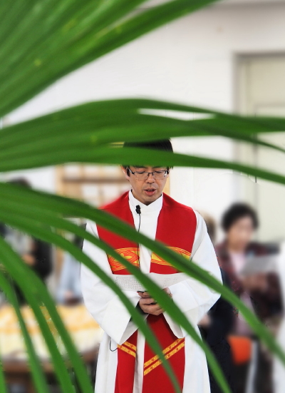 2019年の棕櫚の主日の写真、大きな棕櫚の葉の向こうに司祭の写真