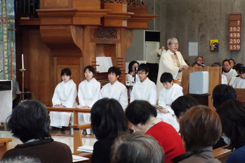 高学年生が祭壇の周りに座っている写真。後ろの説教台では前田司祭が説教しています。